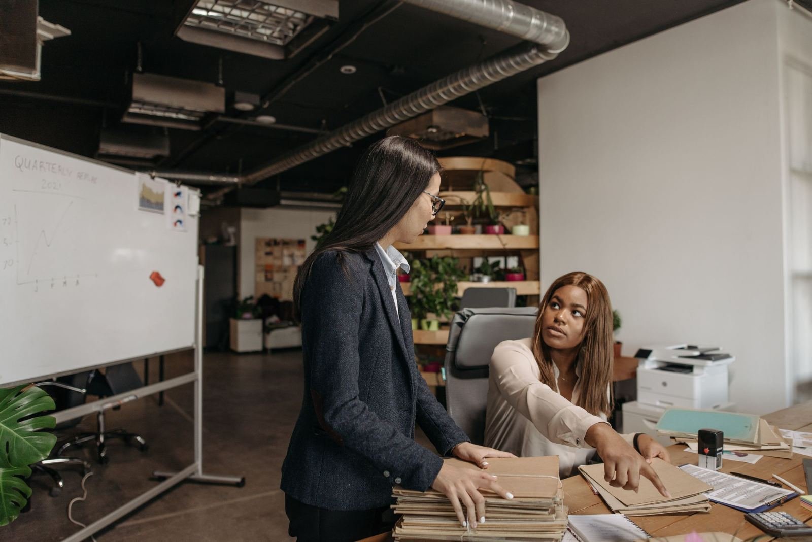 A woman in a blazer discusses paperwork with a seated woman in a bright office