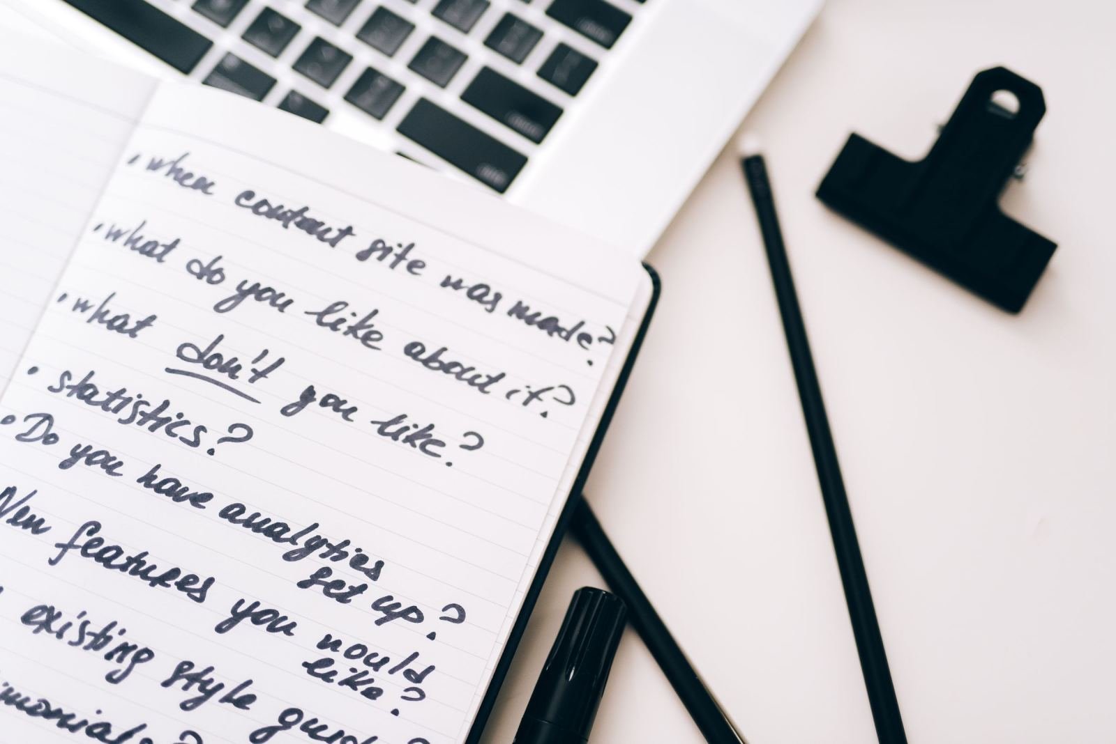 A notebook with handwritten questions next to a laptop and pens on a white desk