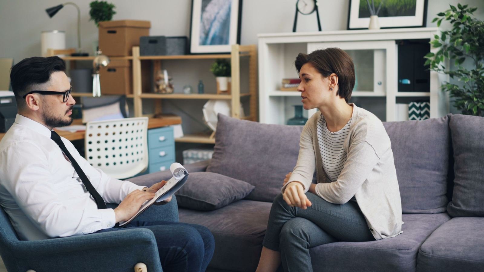 A man in a white shirt and tie talks with a woman sitting on a grey sofa