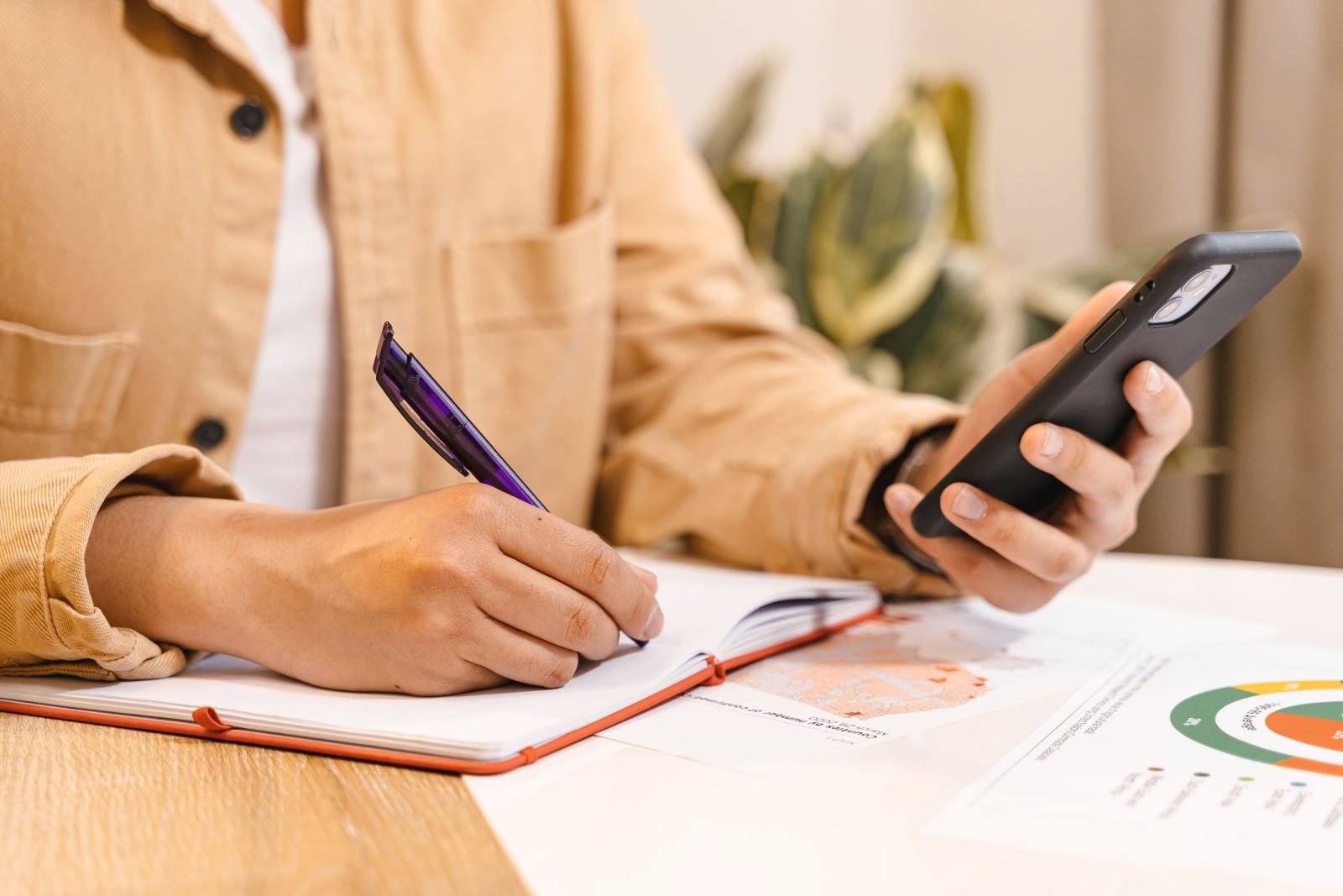 Someone holds a phone in one hand while writing in a notebook on a table