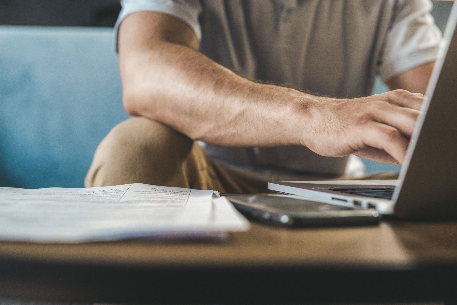 A person typing on a laptop with papers and a smartphone on the desk