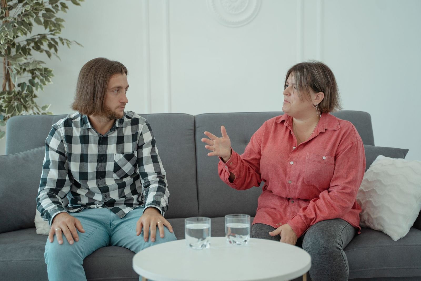 A man and woman sit on a gray sofa and talk to each other