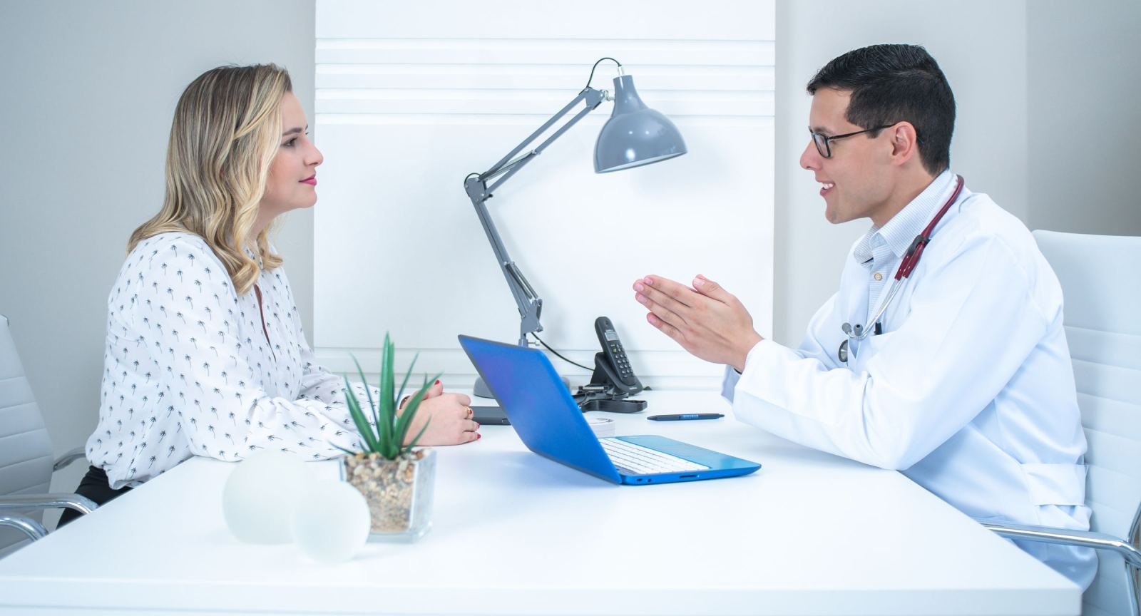 A woman and a doctor in a white coat talk across a desk with a blue laptop
