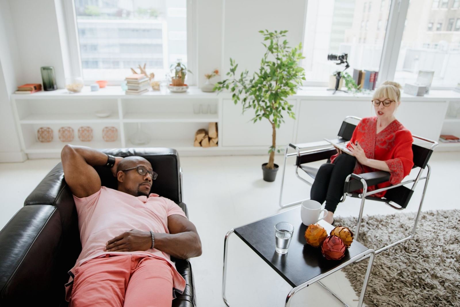 A woman in a red cardigan writes notes while a man leans back in a leather chair.