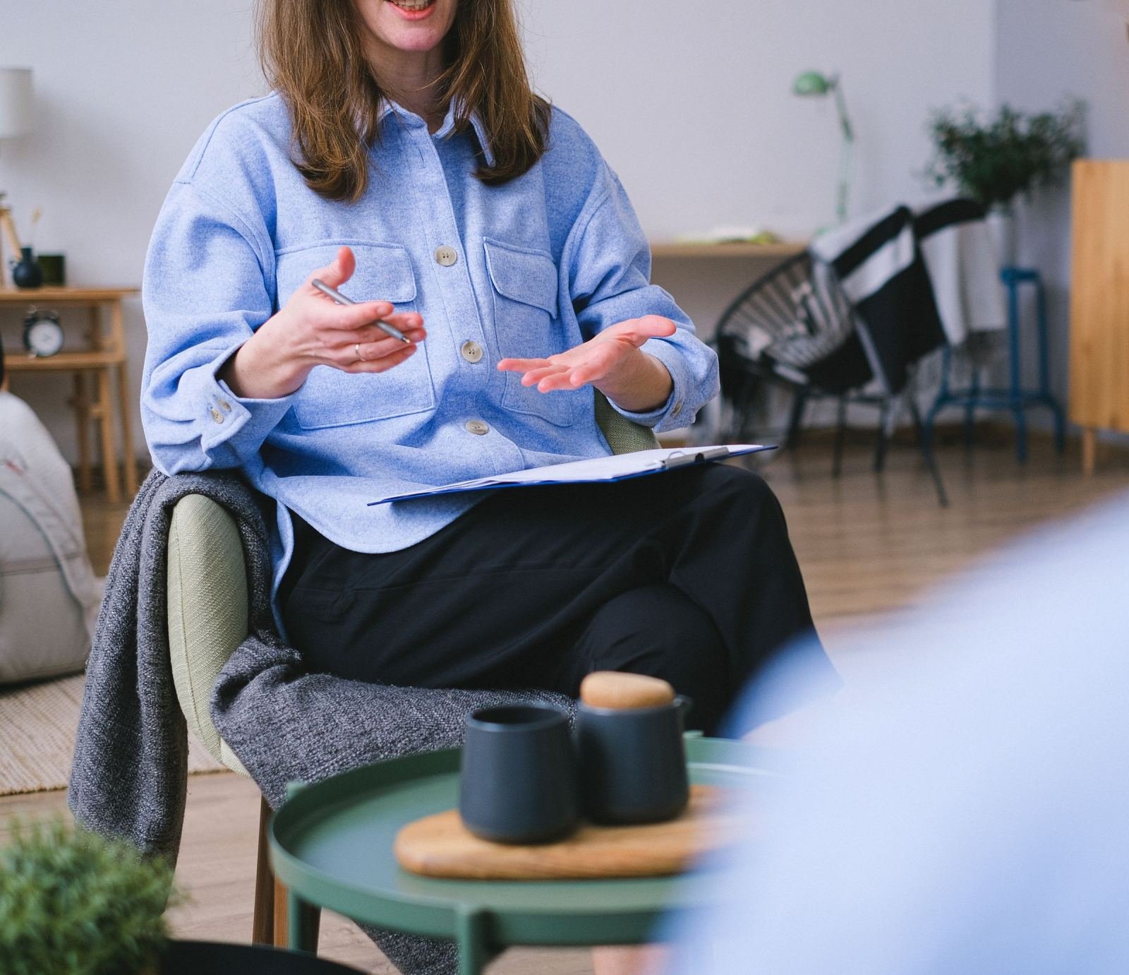 A woman in a blue shirt gestures while holding a pen and clipboard