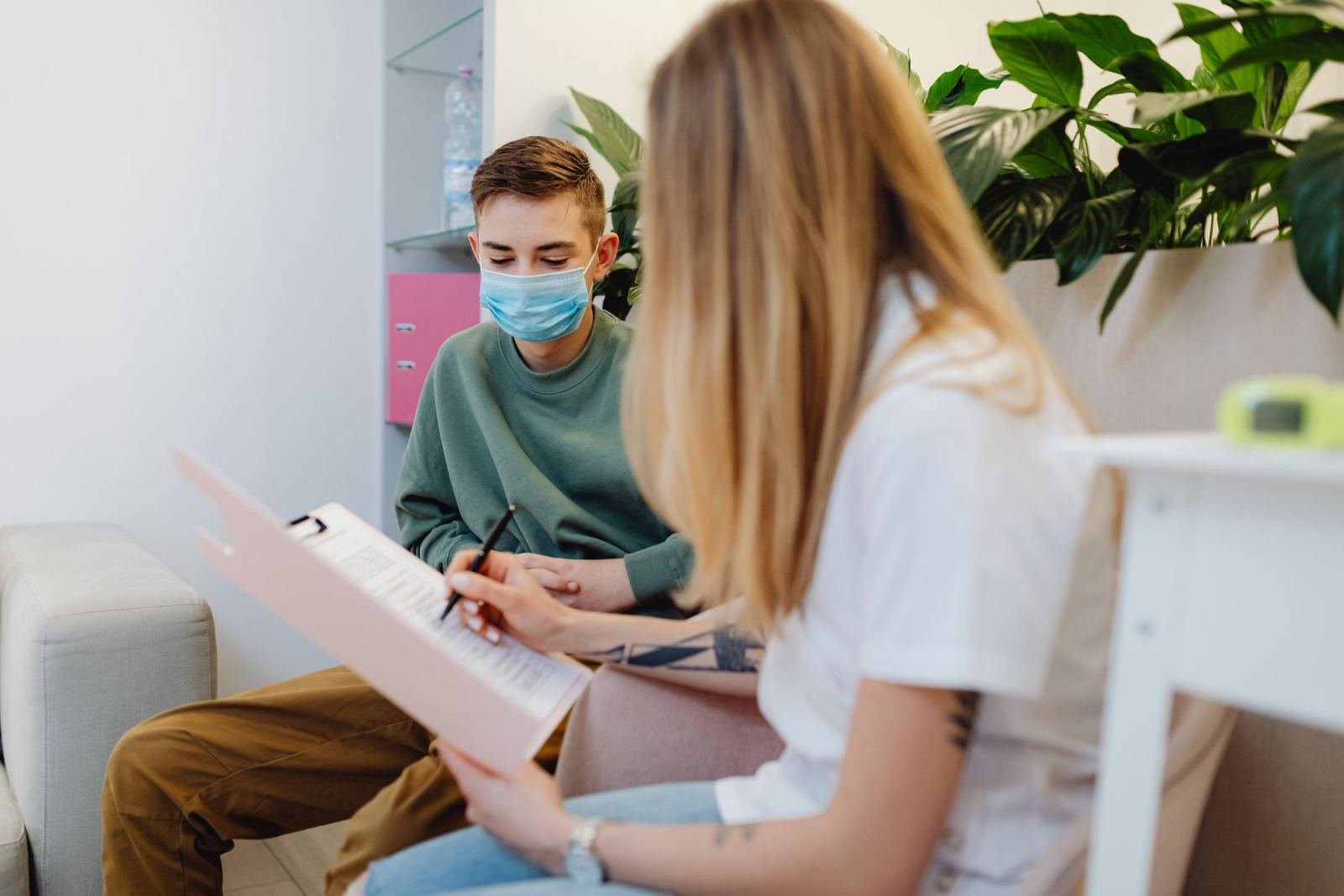 A person in a white shirt writes on a clipboard while talking to a person wearing a mask