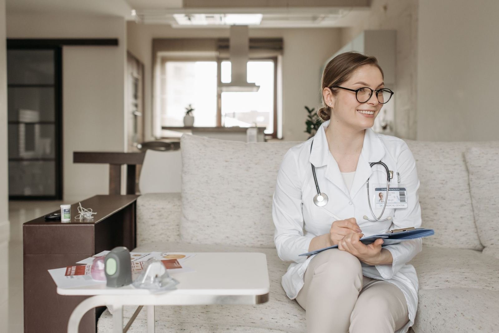 A smiling medical professional in a white coat writes on a clipboard while sitting on a sofa