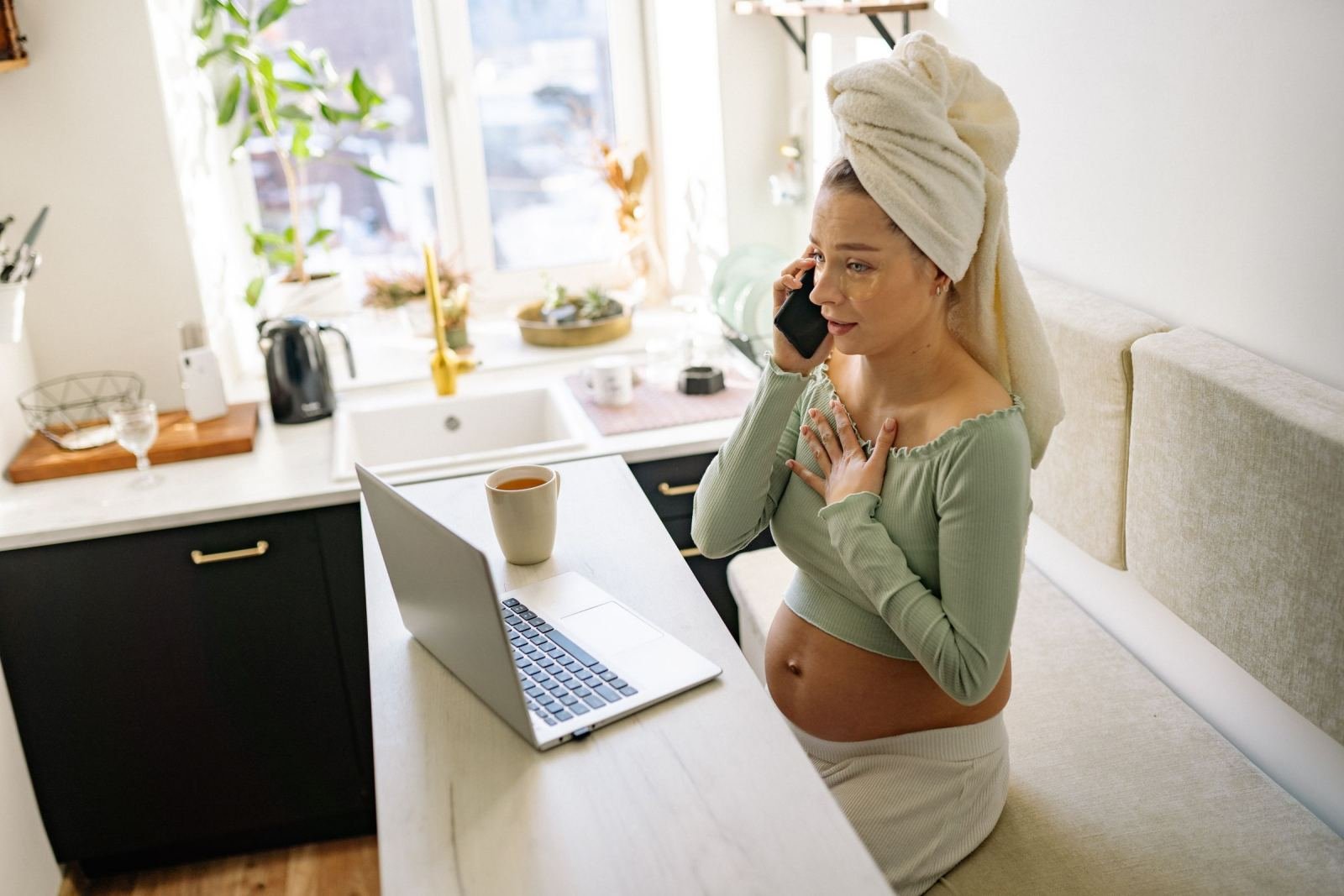A pregnant woman holds a phone to her ear. She stands in her kitchen with a towel wrapped around her head.