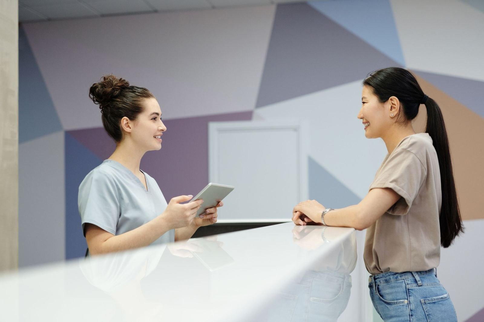 A clinic receptionist with a tablet speaks with a woman at a white front desk