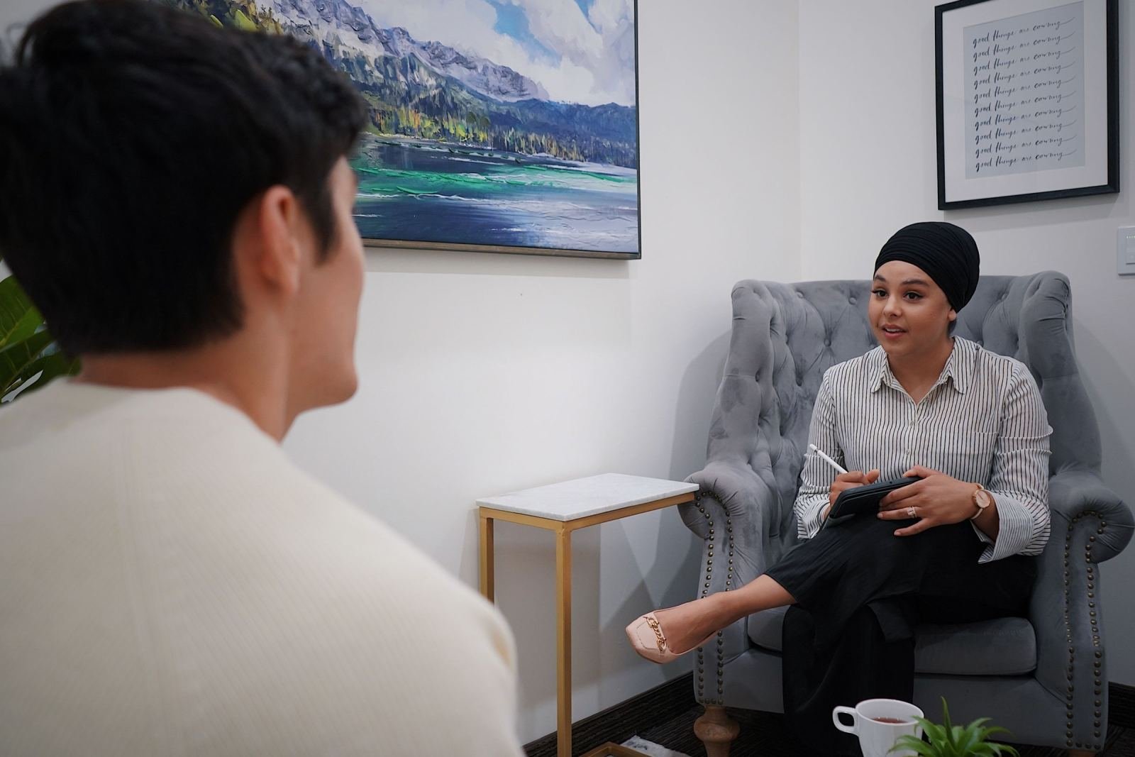 A man talks with a woman wearing a turban while she sits in a gray armchair in a therapy room