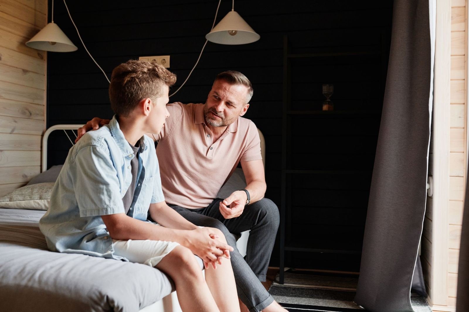 A man rests his hand on a teen boy's shoulder while they talk on a bed.