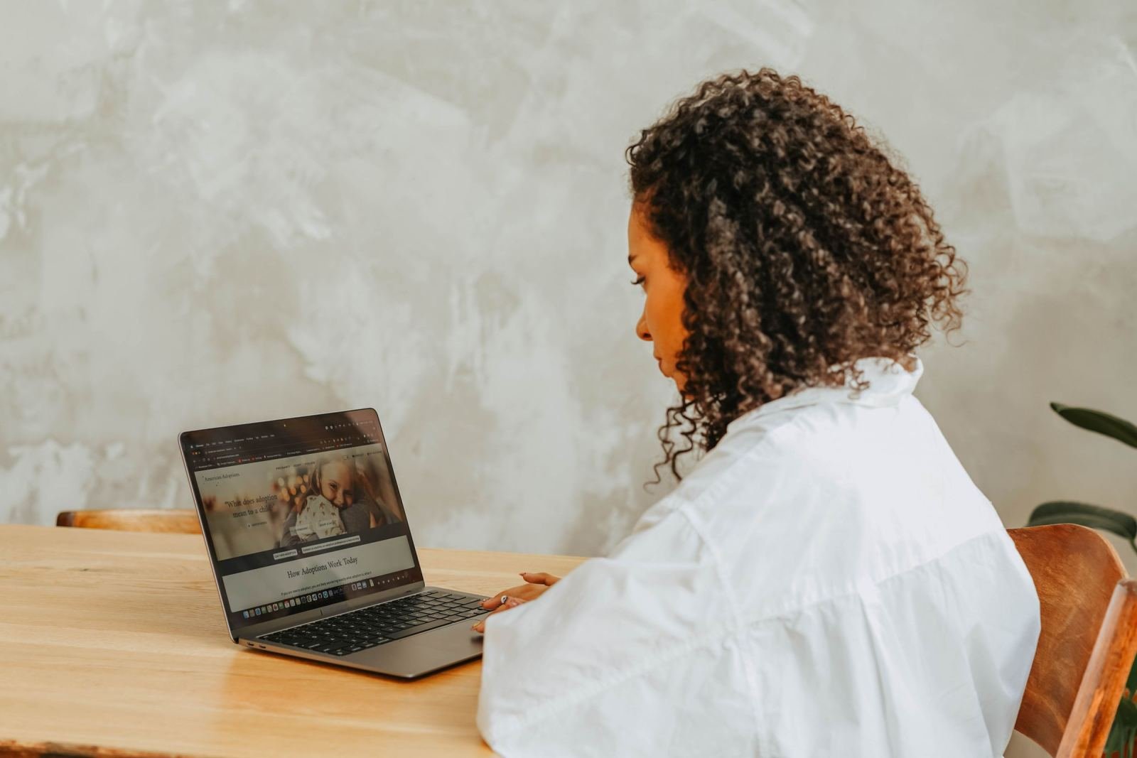 A woman in a white top sits at a wood desk and works on her laptop.