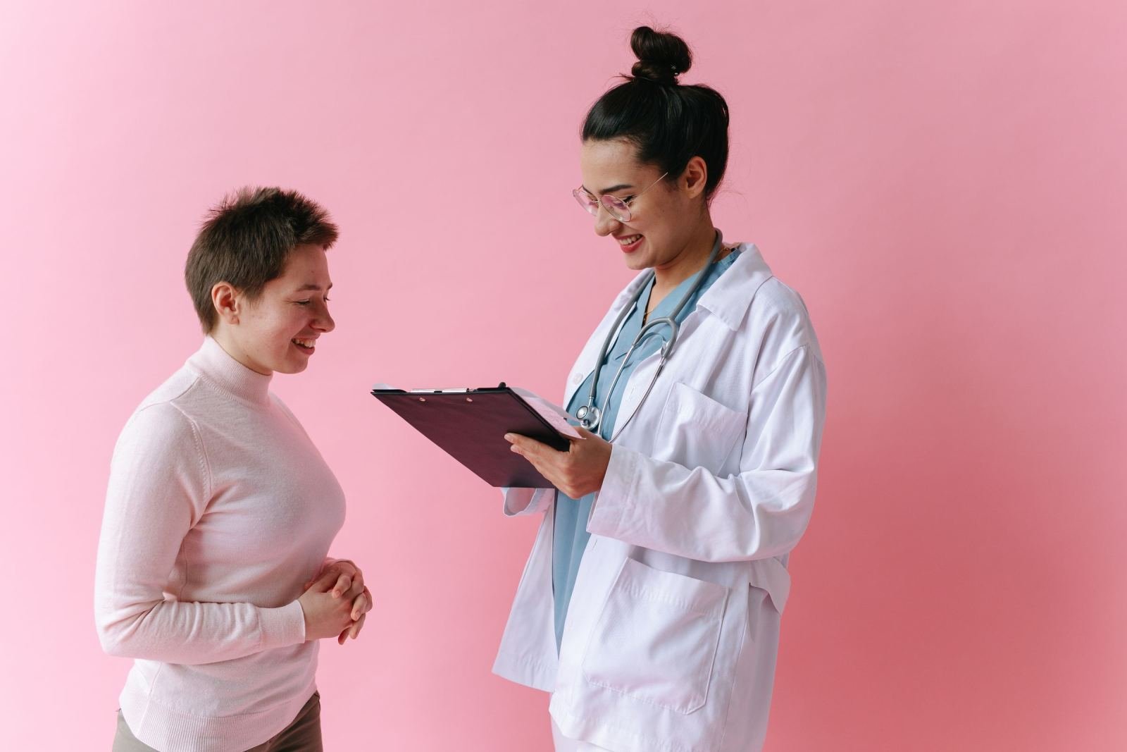 A doctor in a white coat holds a clipboard while smiling at a patient against a pink background