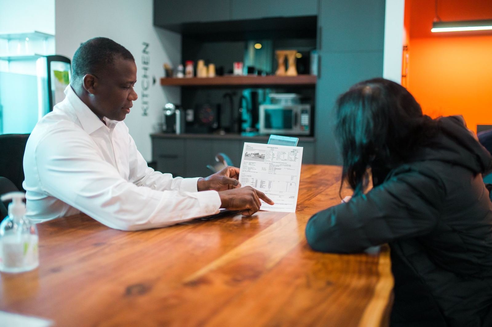 A man in a white shirt points to a document while talking to another person at a wooden table