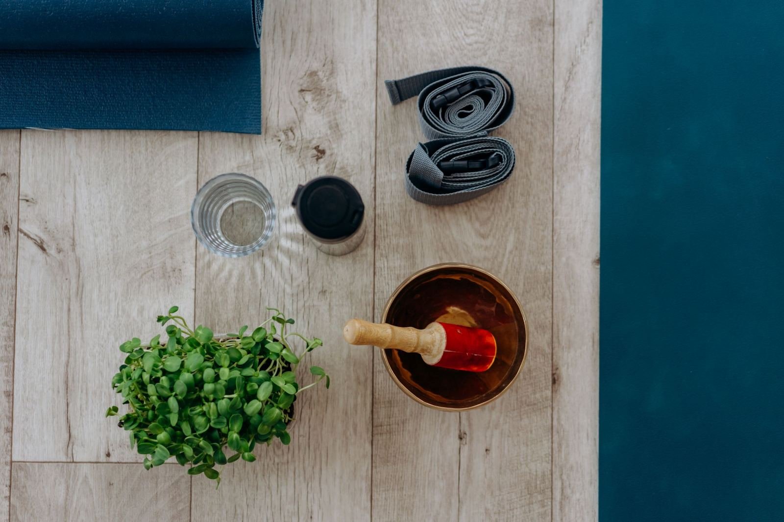 Yoga mat, strap, singing bowl, water, and a plant on a wooden floor