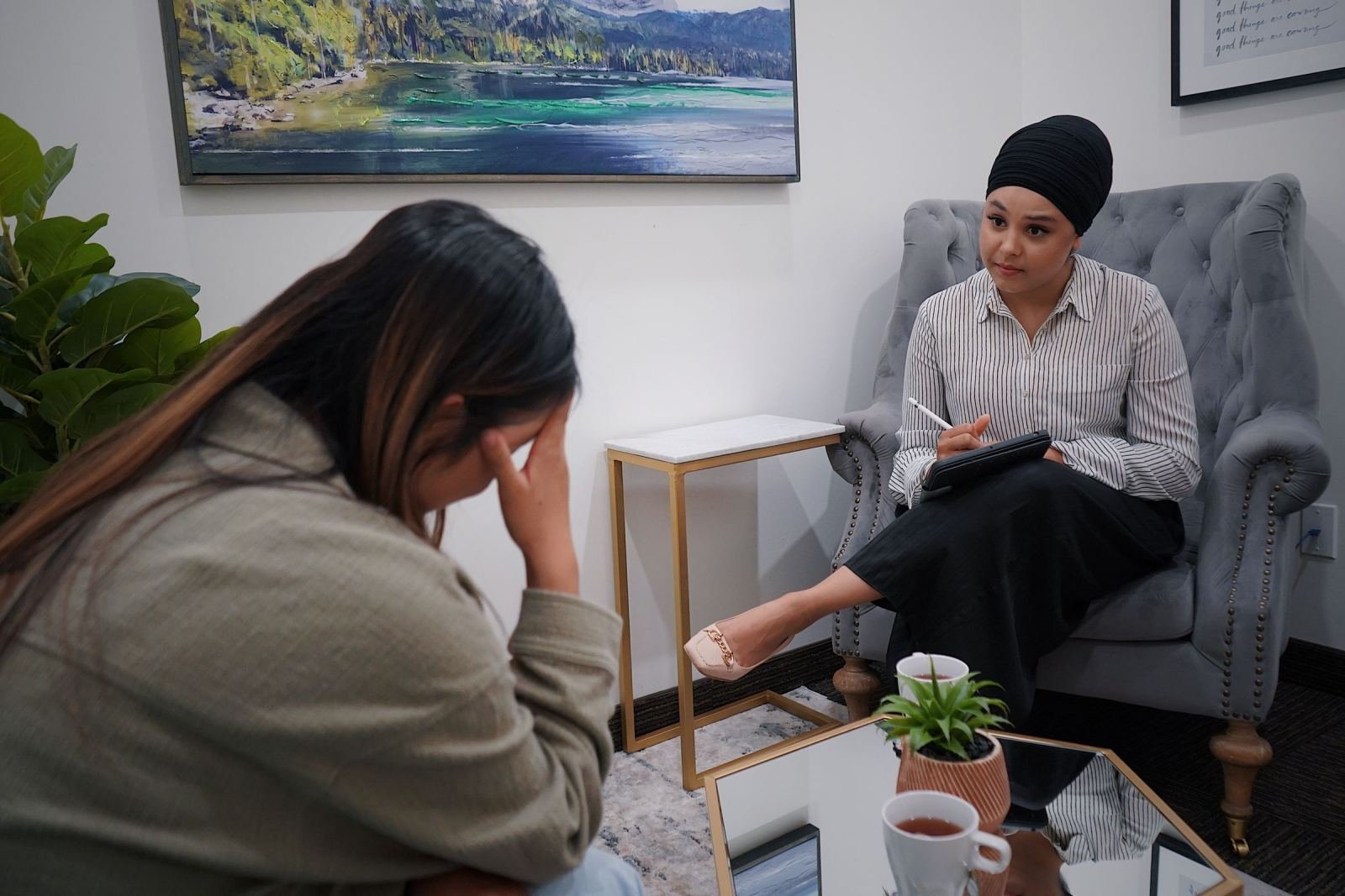 A woman sits in a grey armchair, hand on her forehead, talking to a clinician