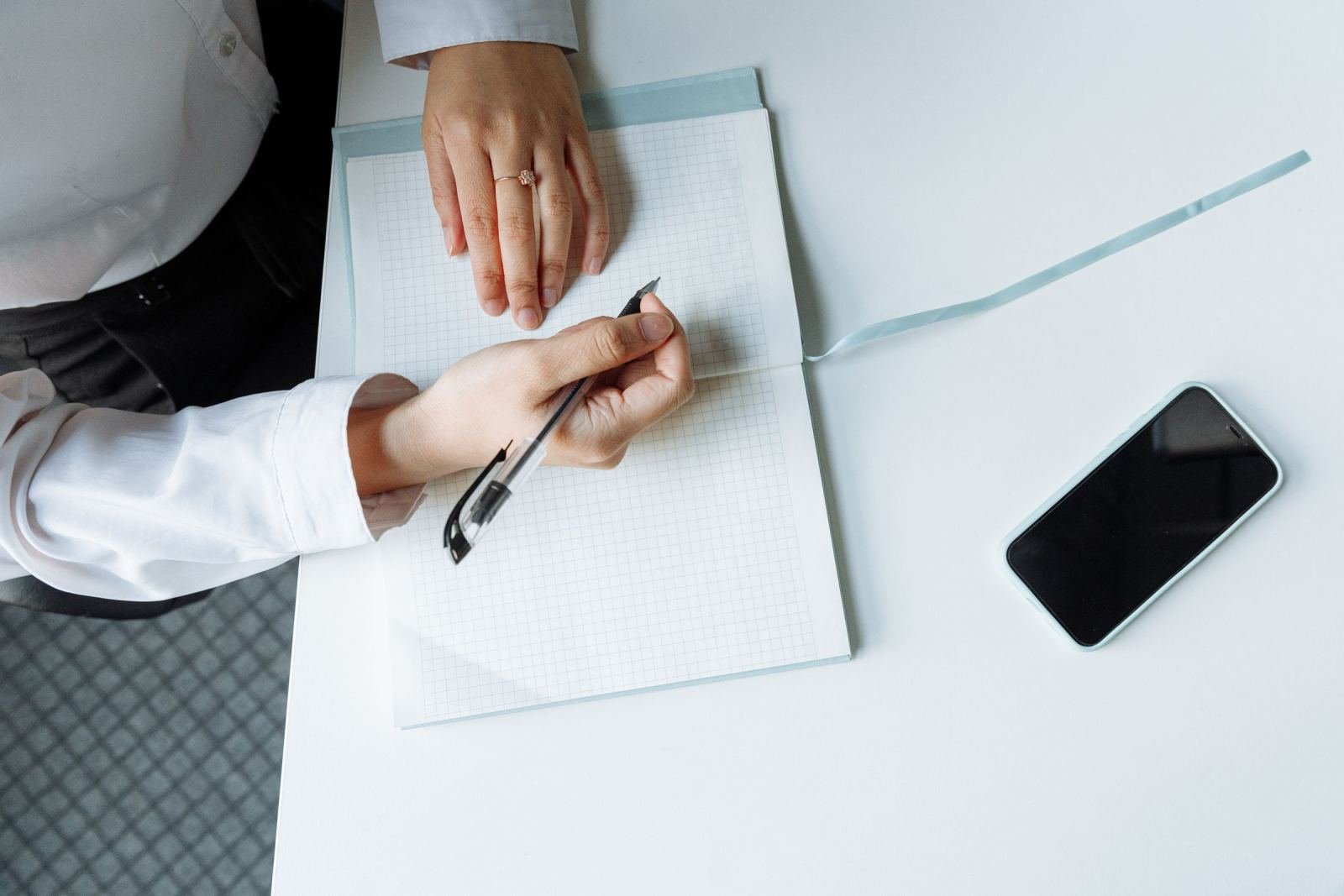 A person in a white shirt writes in a grid notebook next to a smartphone