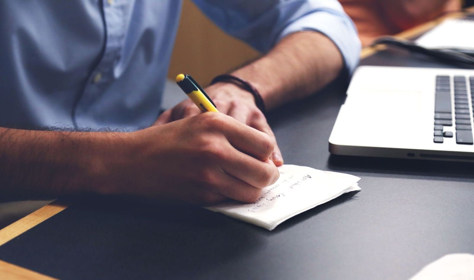 A person writing notes on a piece of paper next to a laptop