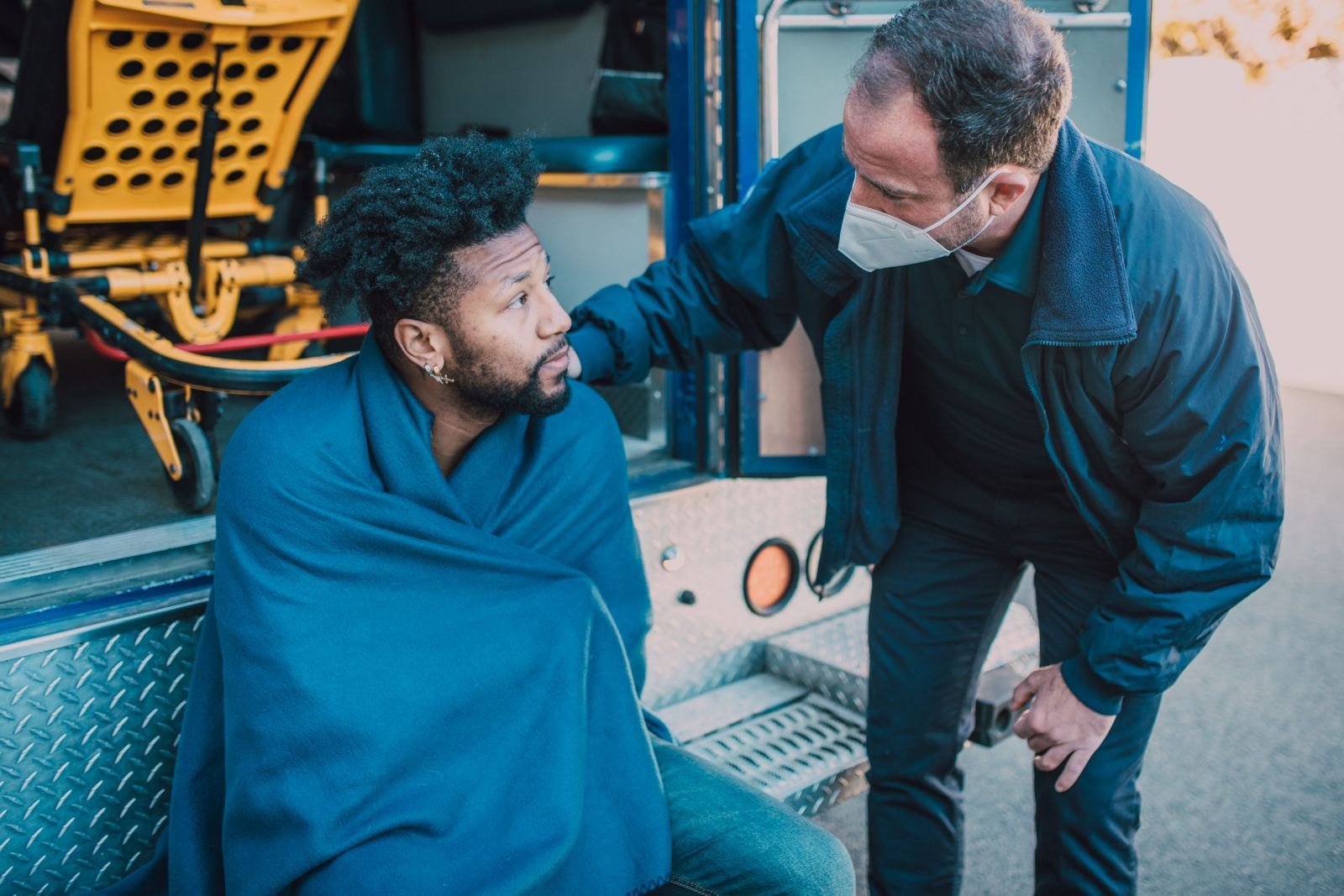 A paramedic in a mask comforts a man wrapped in a blue blanket in an ambulance
