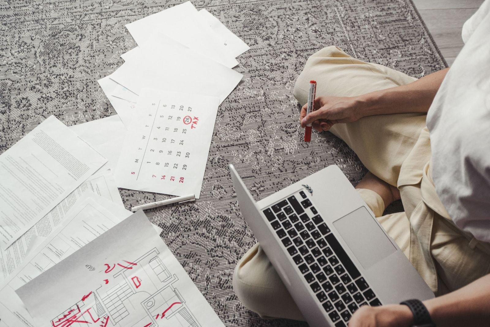 A person sits on a rug with a laptop and scattered papers, including a calendar