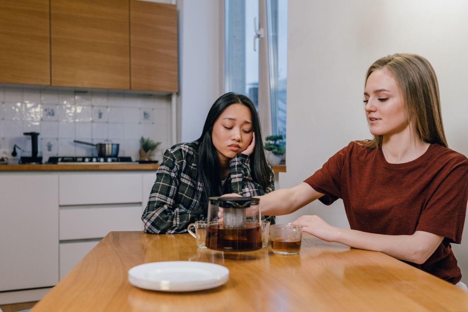 Two women sit at a kitchen table with a teapot, one listening while the other speaks