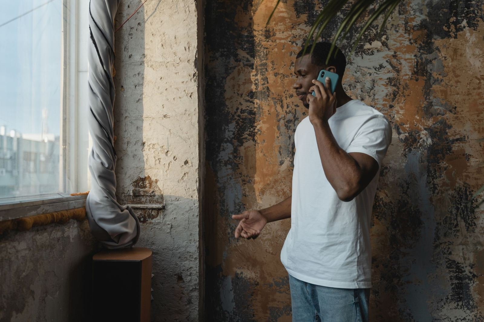 A man in a white t-shirt talks on a blue smartphone while standing by a textured wall