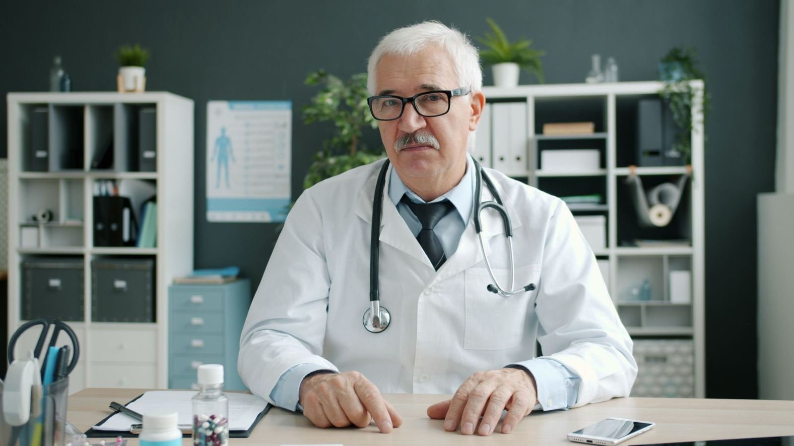 A doctor in a white lab coat and stethoscope sits at his desk