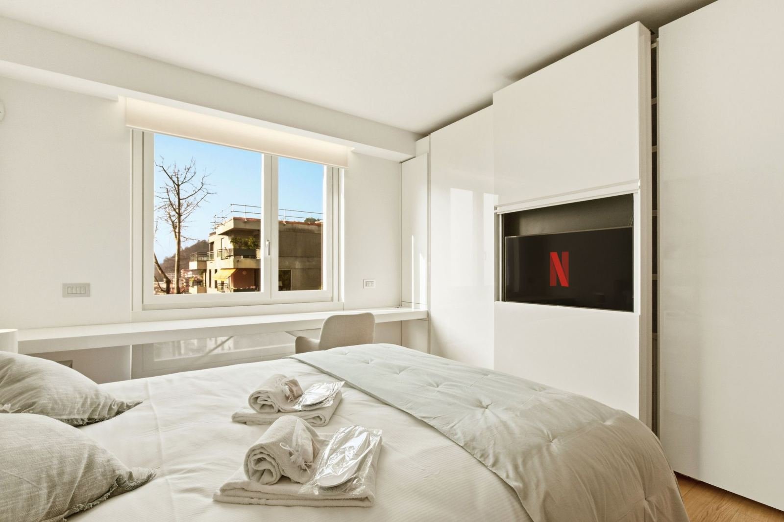 A bright, modern bedroom with white linens and a television built into the wall