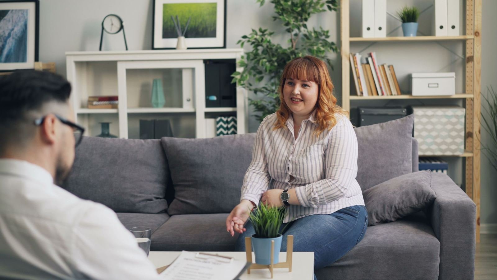 A woman sits on a gray sofa talking to a man in a bright office