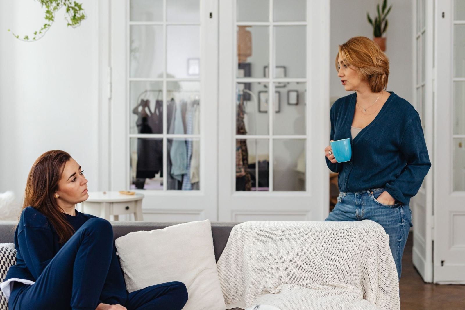 A woman sits on a couch while another stands holding a cup. They talk in a bright living room.