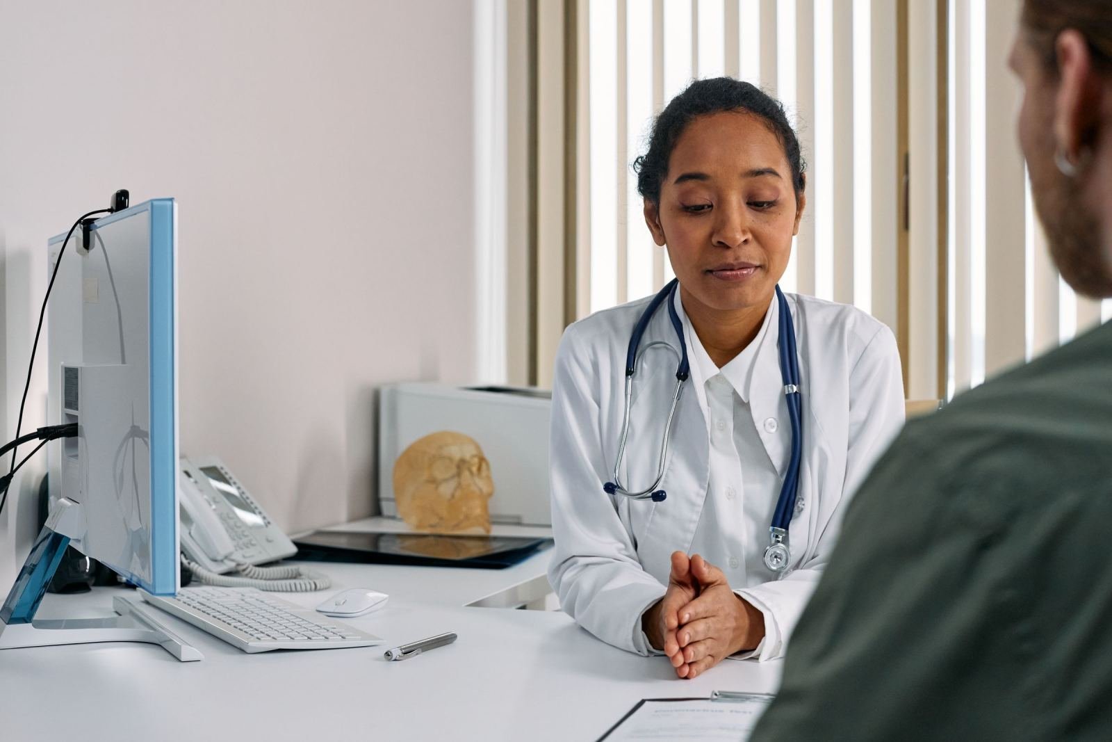A clinician in a white coat and stethoscope talks with a patient in a bright office