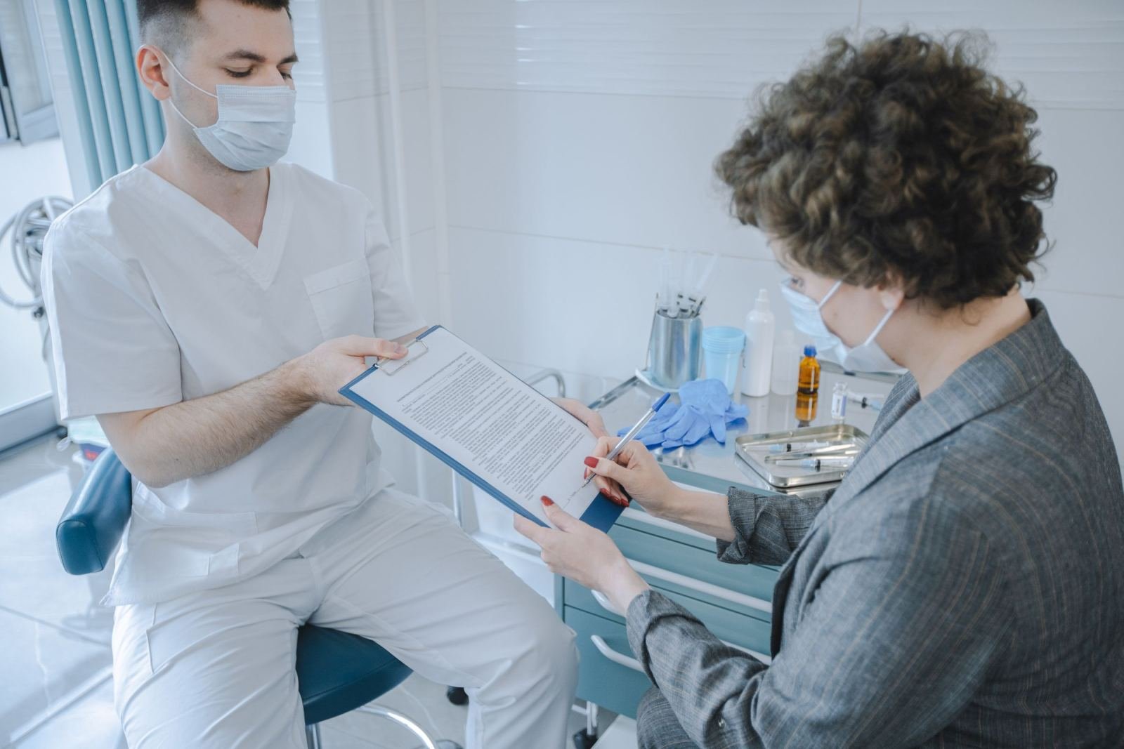 A healthcare worker in a mask holds a clipboard for a woman to sign in a clinic