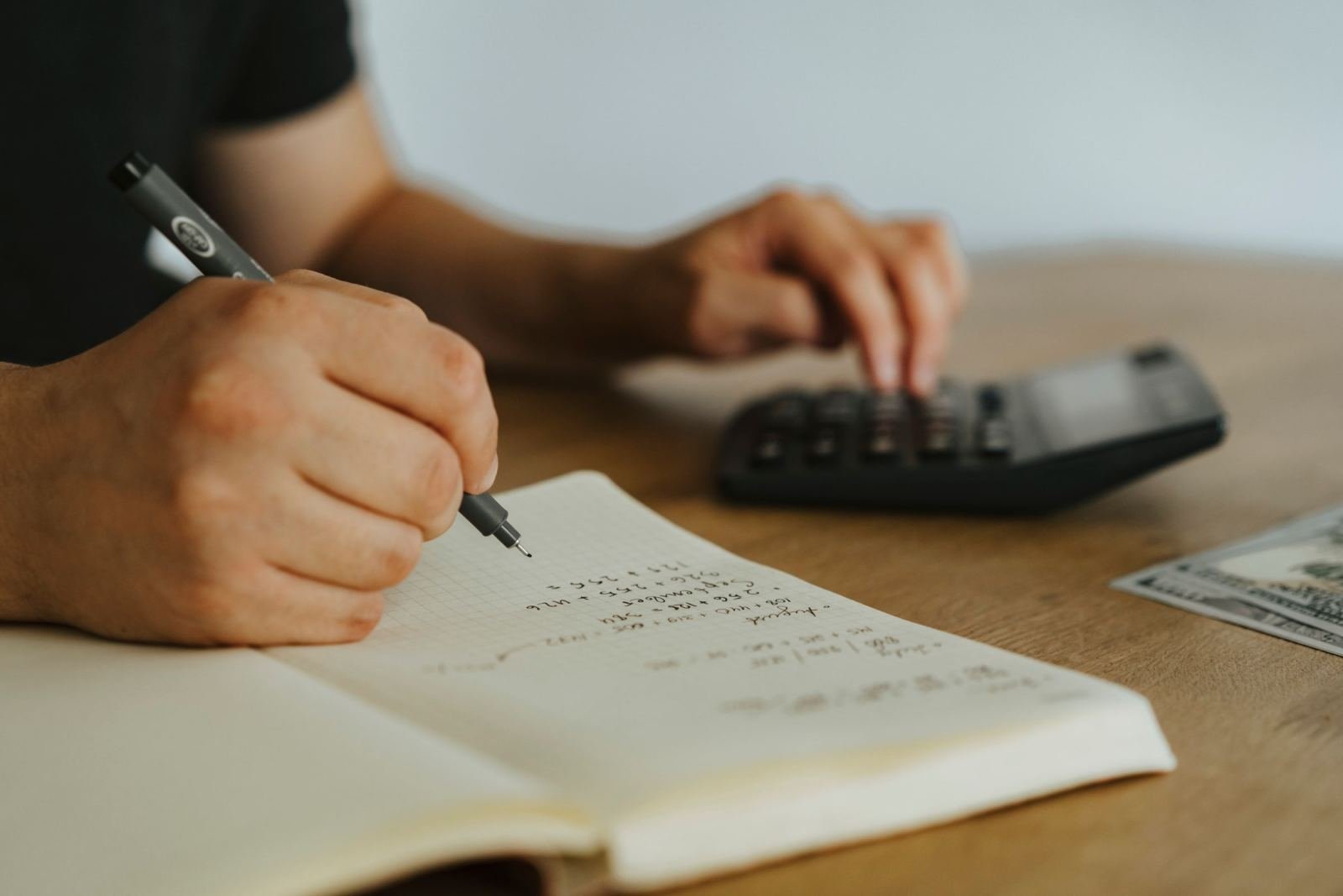 A person using a calculator and pen to write notes in a notebook next to cash