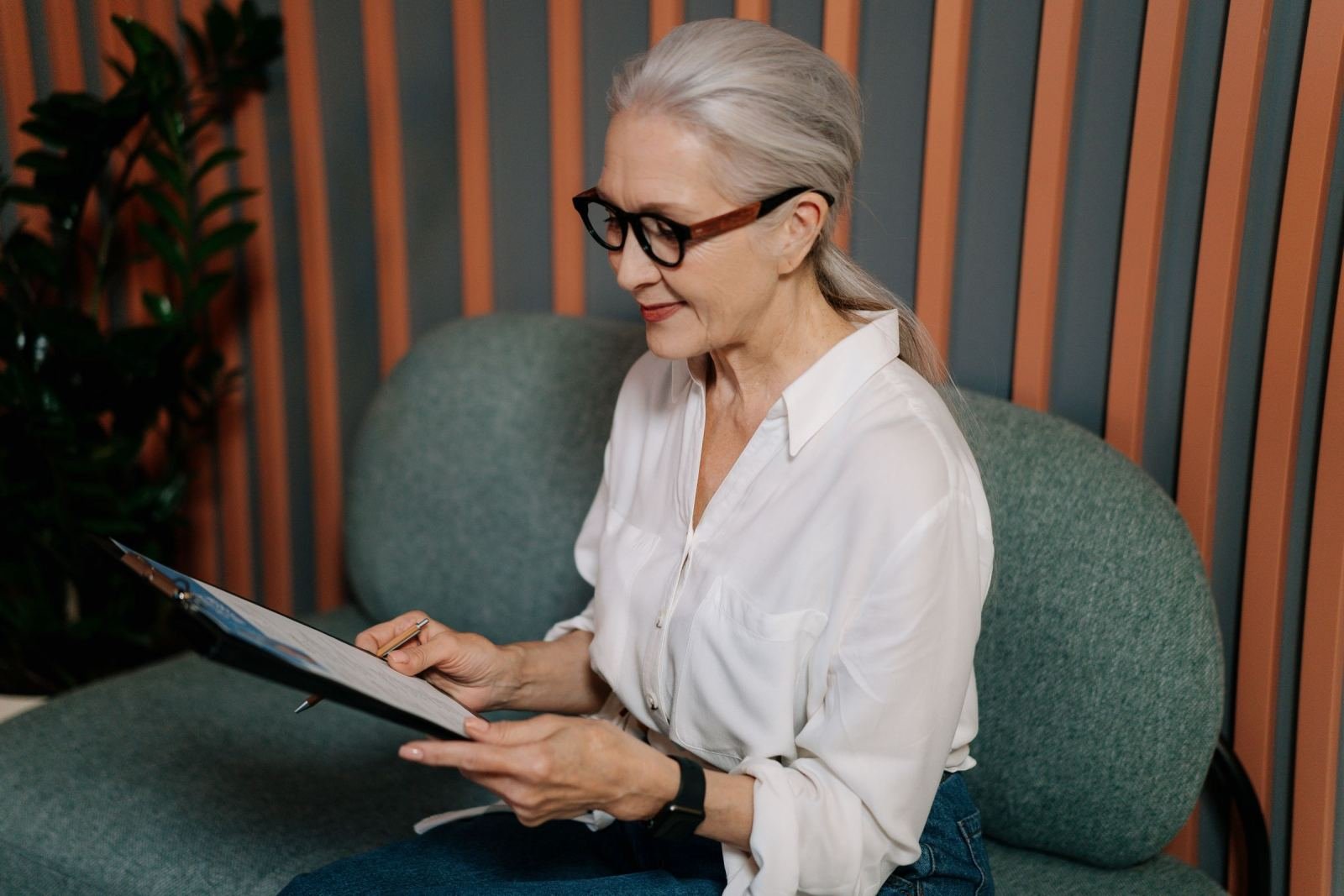 A woman with glasses writes on a clipboard while sitting on a green sofa