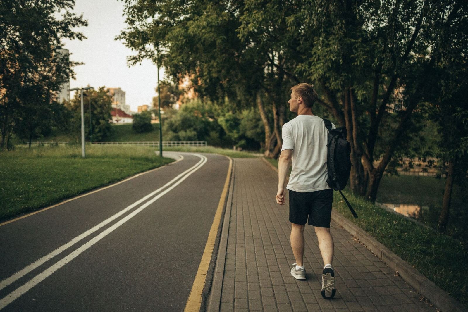 A man carrying a backpack moves along a paved trail in a green park.