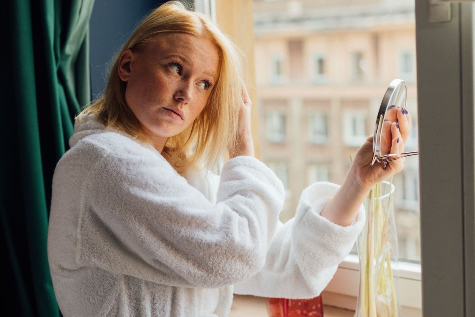 A woman in a white bathrobe looks at her reflection in a handheld mirror