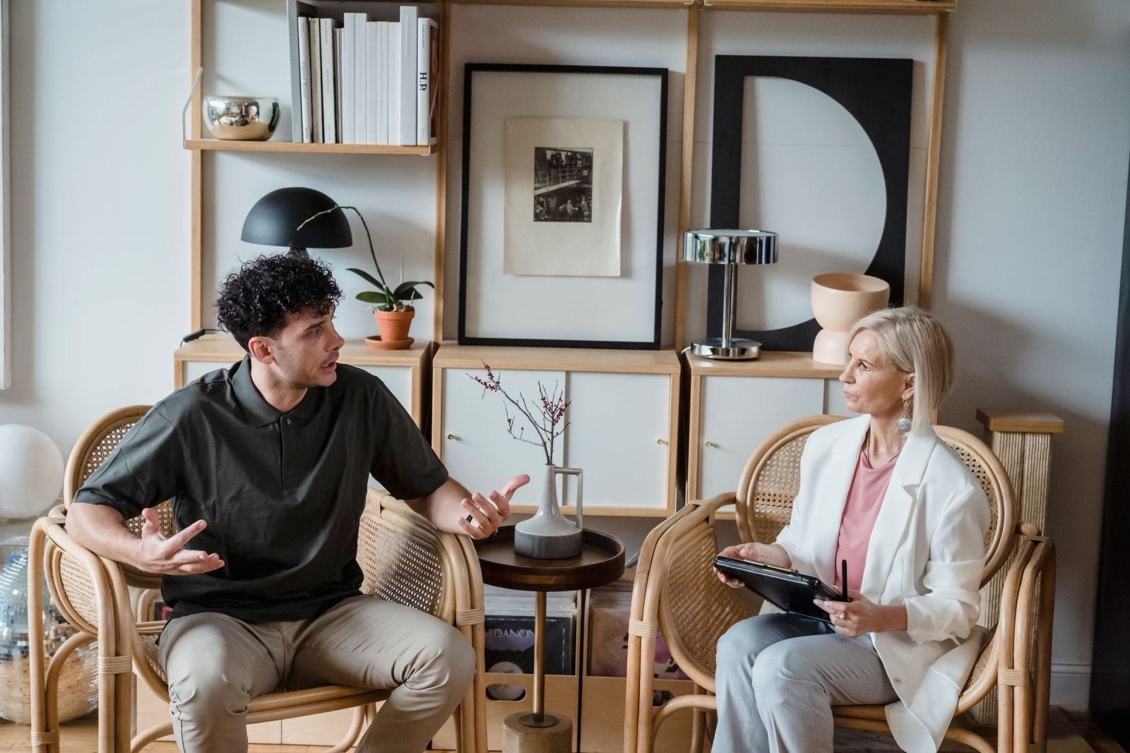 A man and a woman sit in a bright office with wicker chairs and talk