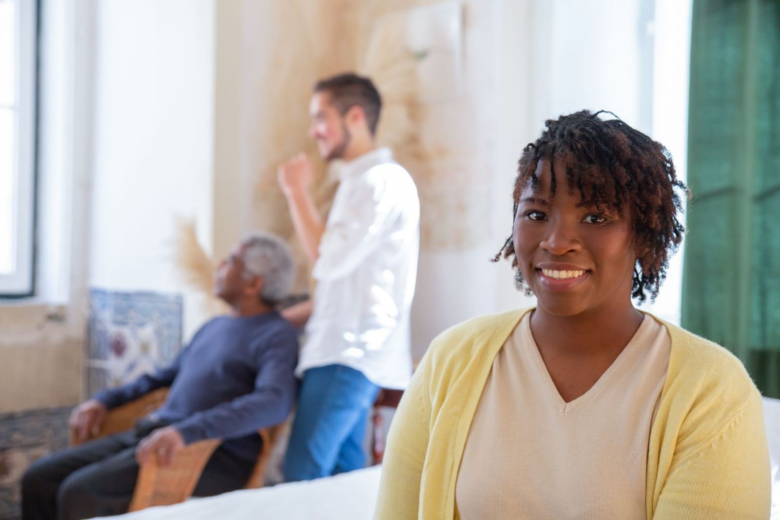 A smiling woman in a yellow cardigan with two men talking in the background