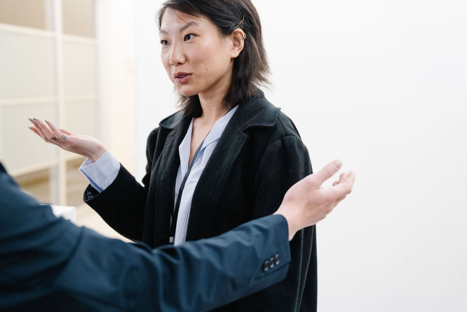 A woman in a black coat gestures with her hands while talking to another person