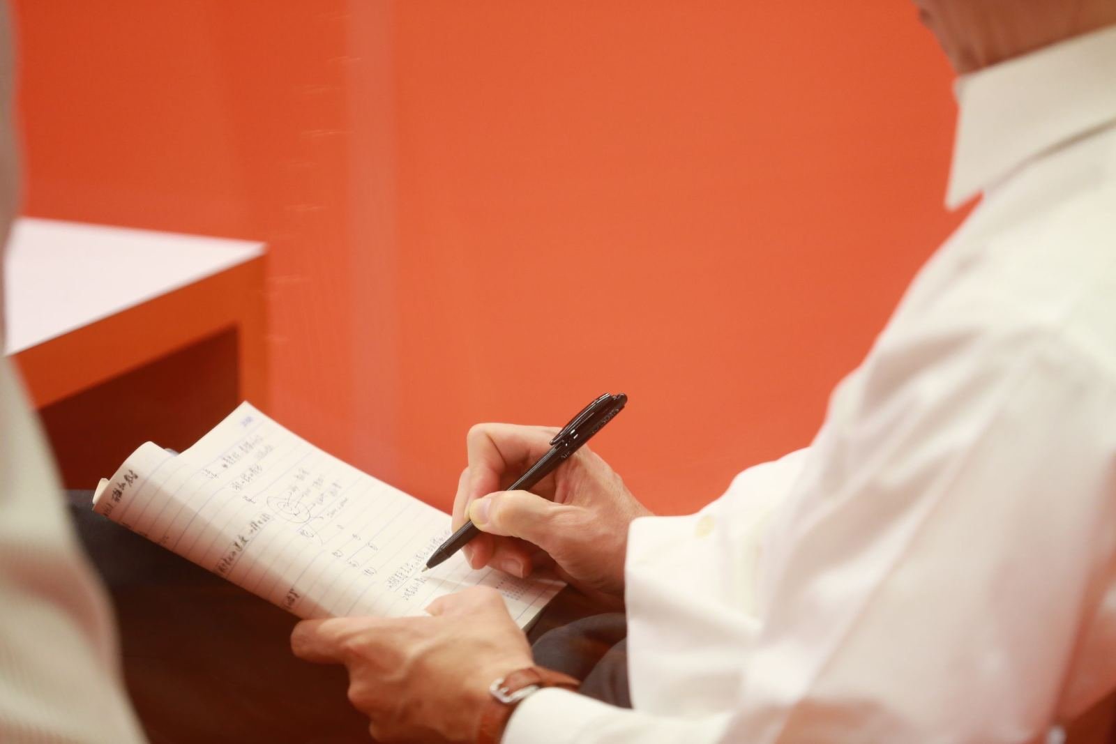 A person in a white shirt writing in a notebook with a black pen