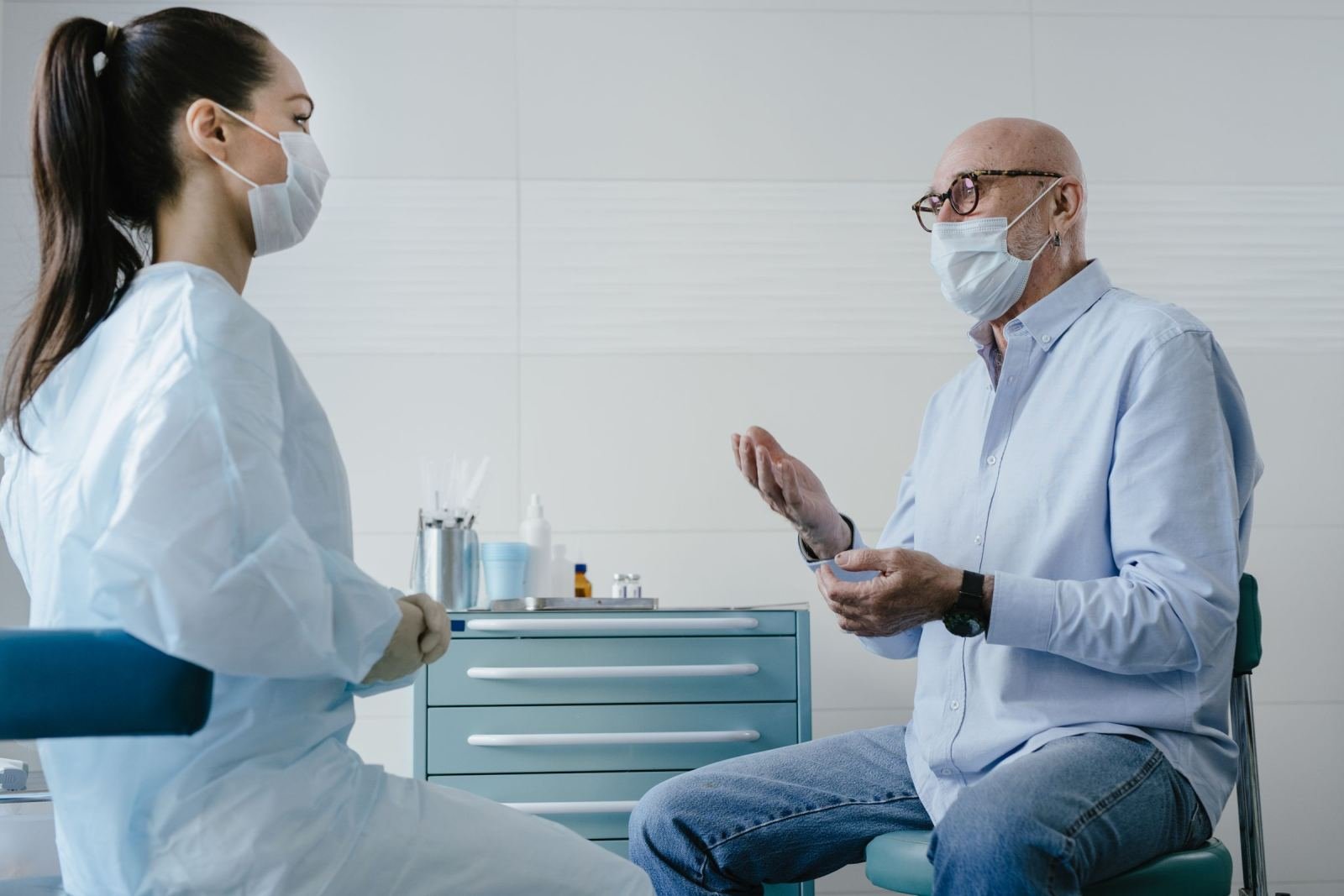 A medical professional in a gown and mask speaks with a patient in a clinical setting
