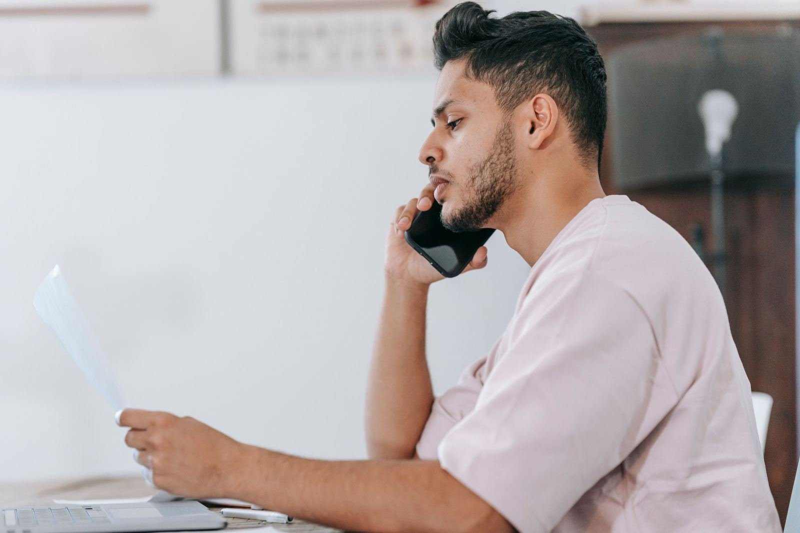 A man on a phone call looks at a document while sitting at a desk with a laptop