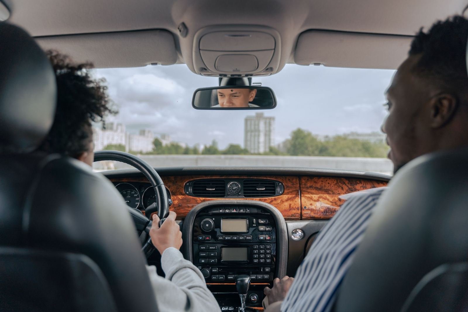 Two people sit in the front seats of a car, with the driver's eyes visible in the rearview mirror