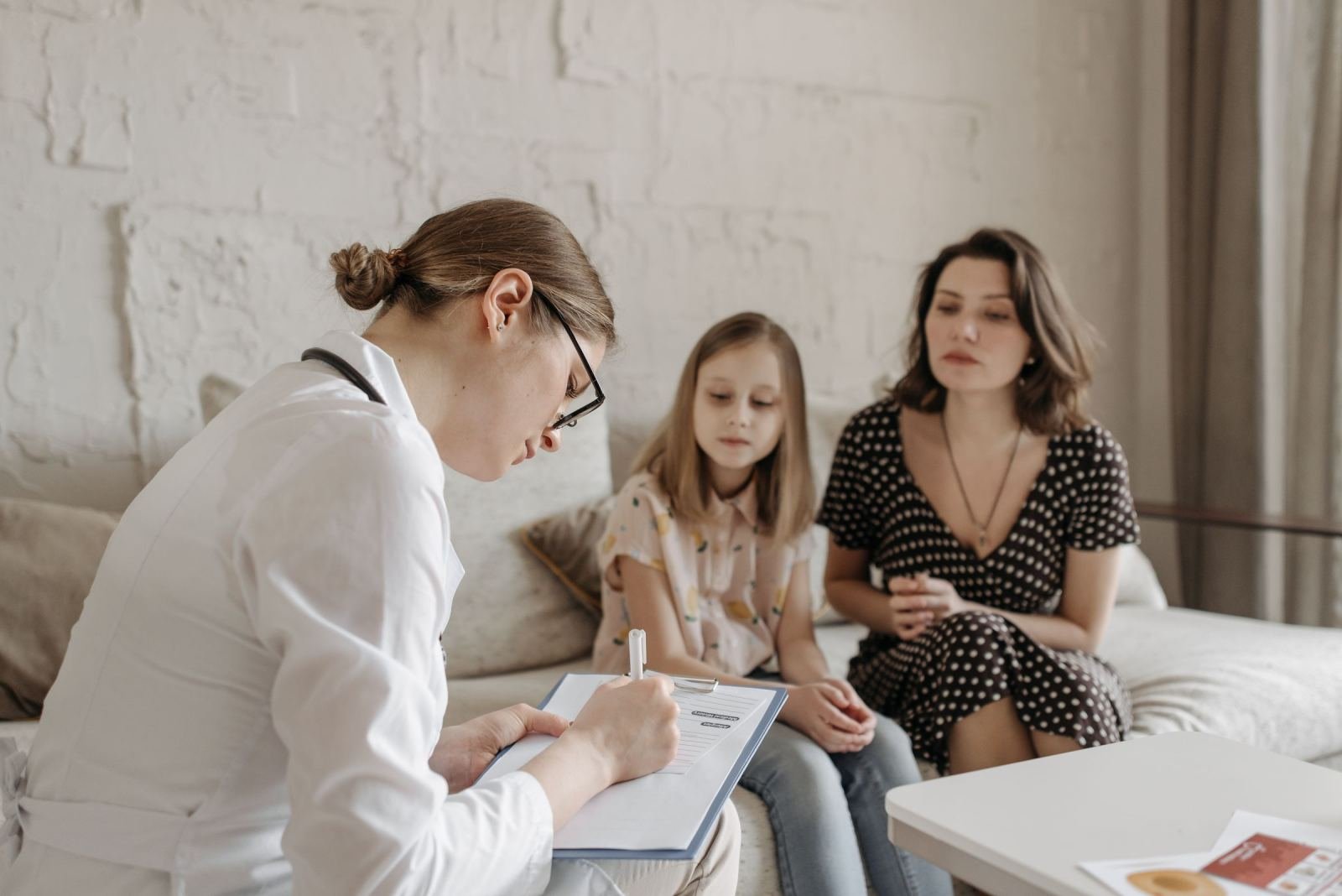 A clinician in a white coat writes on a clipboard while talking to a woman and child