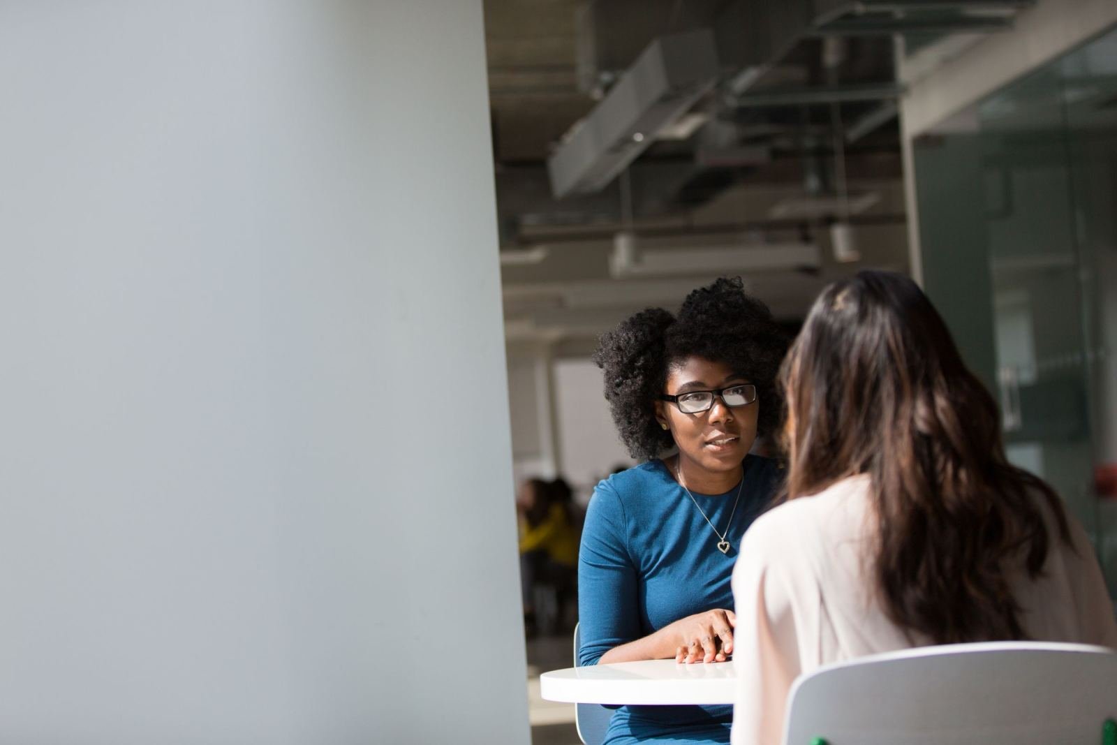 Two women sit at a white table in a modern office having a conversation