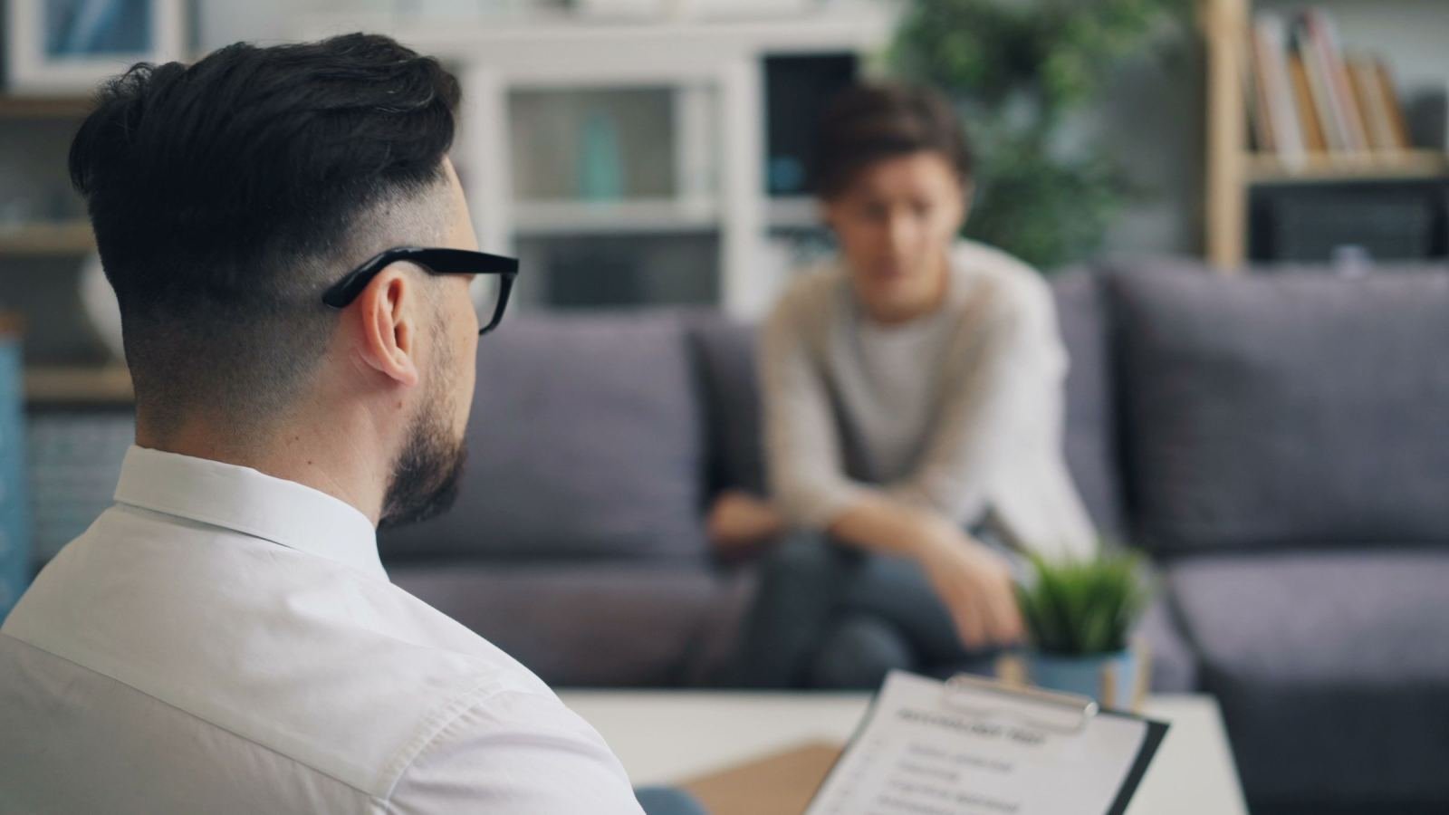 A person sits on a gray sofa while a counselor holds a clipboard and talks to them.