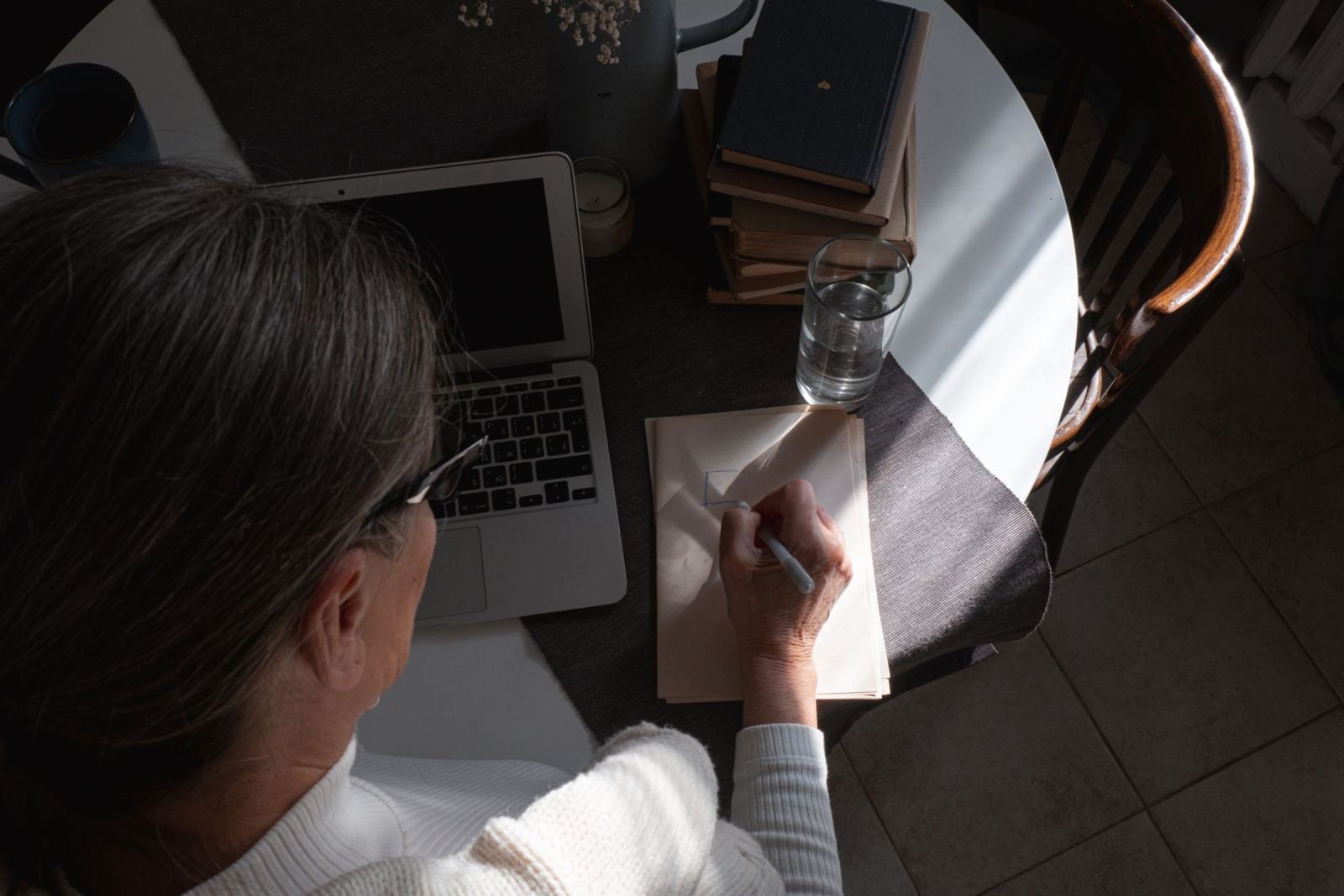 A person with gray hair writes in a notebook at a table with a laptop and books
