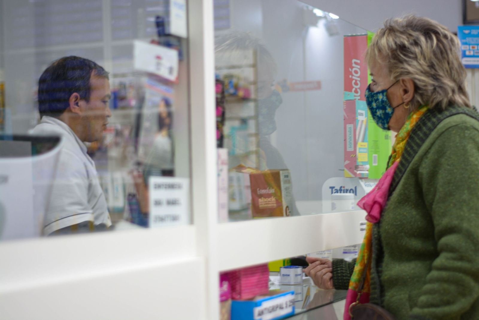 A masked woman speaks with a pharmacist across a glass divider.