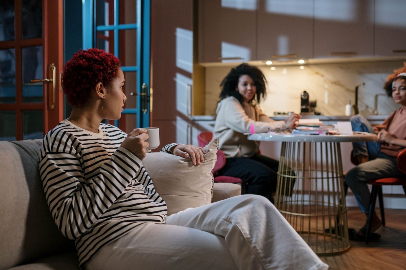 A woman with a mug sits on a sofa while two other women talk at a table in the background
