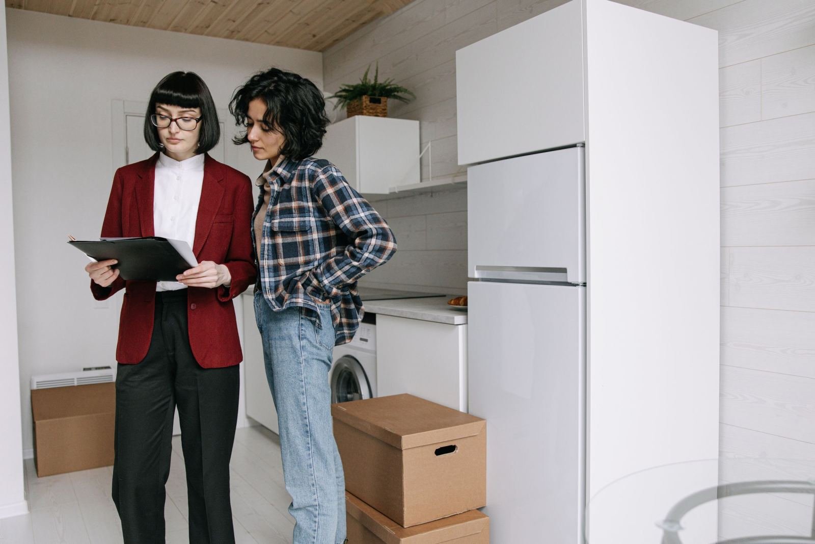 Two women look at a clipboard in a bright kitchen with cardboard boxes on the floor